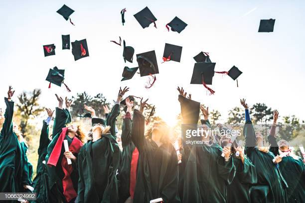 Large group of happy college students celebrating their graduation day outdoors while throwing their caps up in the air.